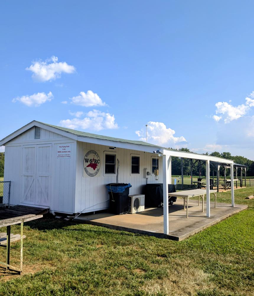 White clubhouse with covered patio on a sunny day, surrounded by green grass.