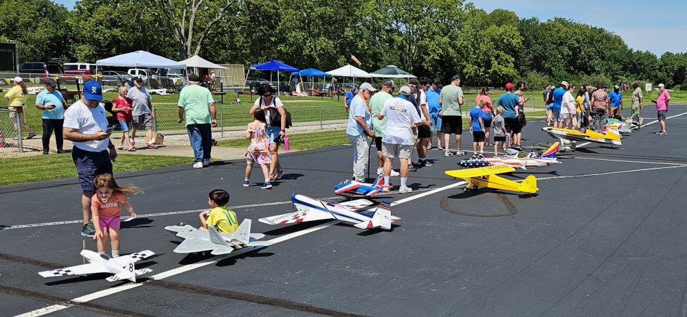 People and children viewing model airplanes on a sunny runway.
