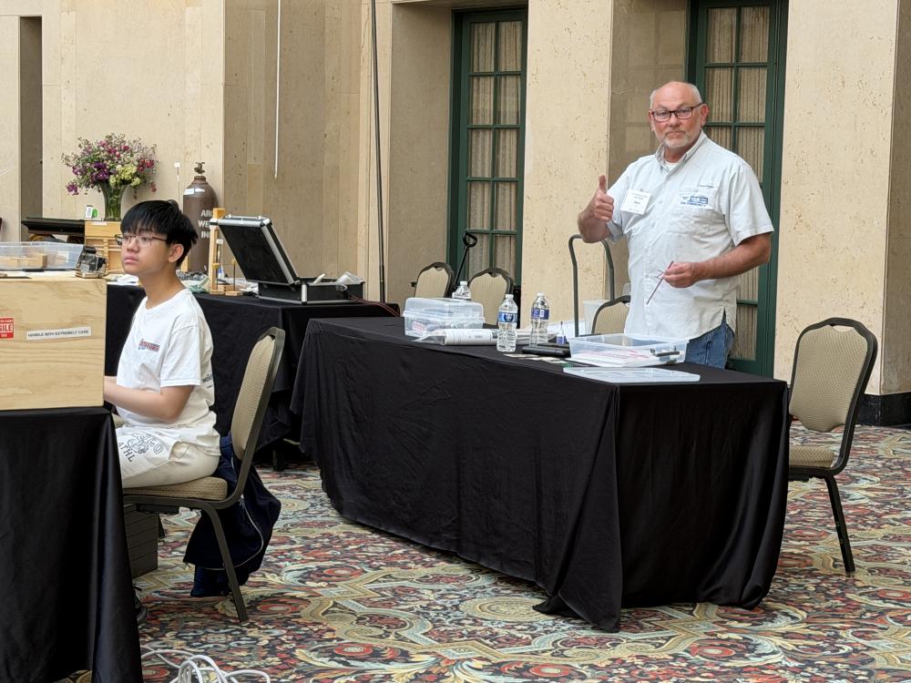 Two people seated at tables in a large hall, one giving a thumbs-up.