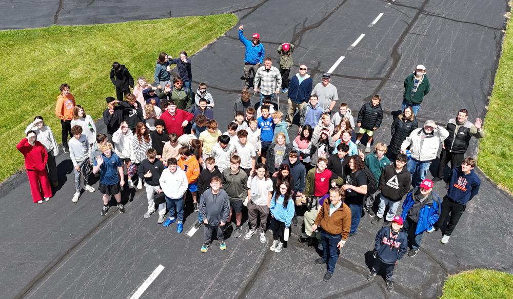 Aerial view of a large group posing on a tarmac, with grass on the sides.