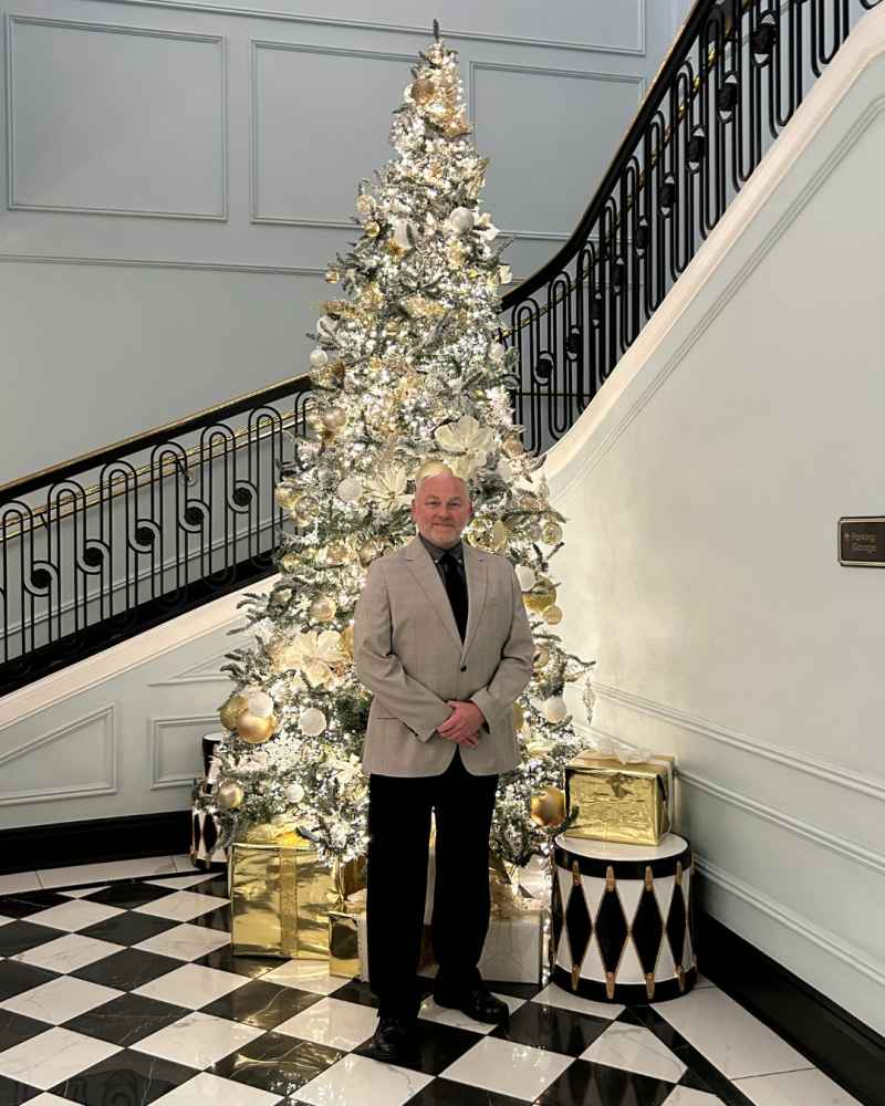 Man in a beige blazer stands by a decorated Christmas tree in a grand staircase setting.