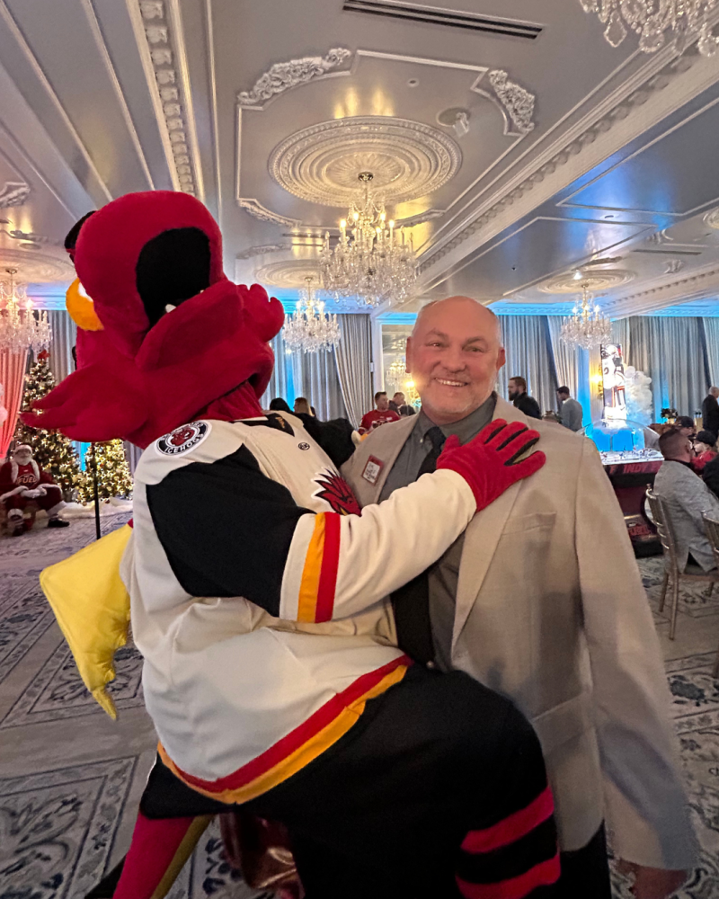 Mascot playfully embraces smiling man in elegant ballroom.