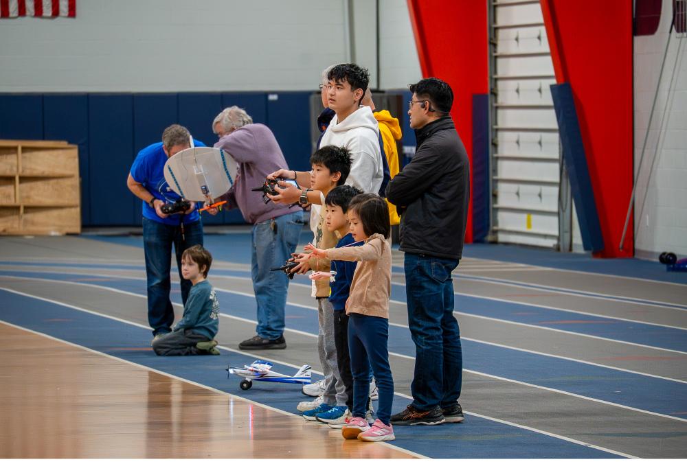 People observing and controlling drones indoors, adults and children engaged.