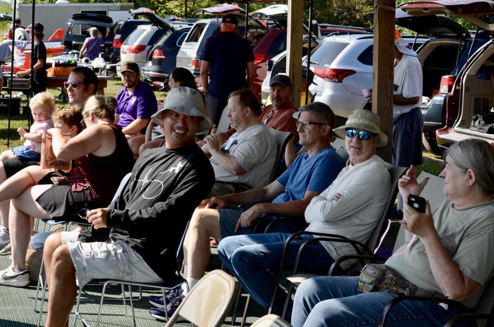 People sitting under a canopy at an outdoor event, some smiling, others using phones.