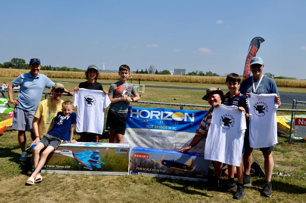 Group celebrating with RC planes and banners outdoors on a sunny day.