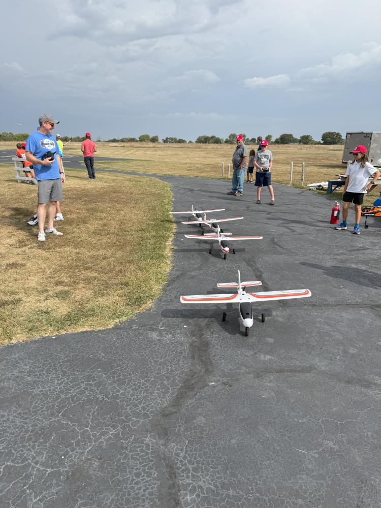 Remote-controlled planes on tarmac, people standing nearby on grassy field.