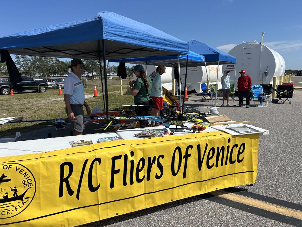 Outdoor booth with people under tents, banner reads "R/C Fliers of Venice."