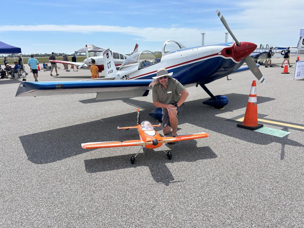Man squatting next to a small orange model plane on an airfield.