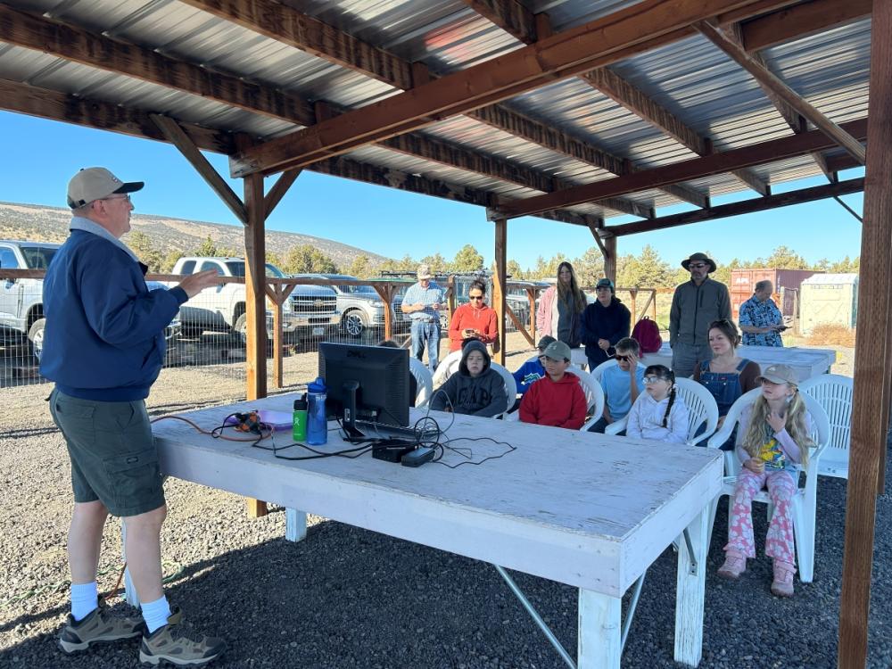 Outdoor class with children seated, instructor at a table with a monitor.