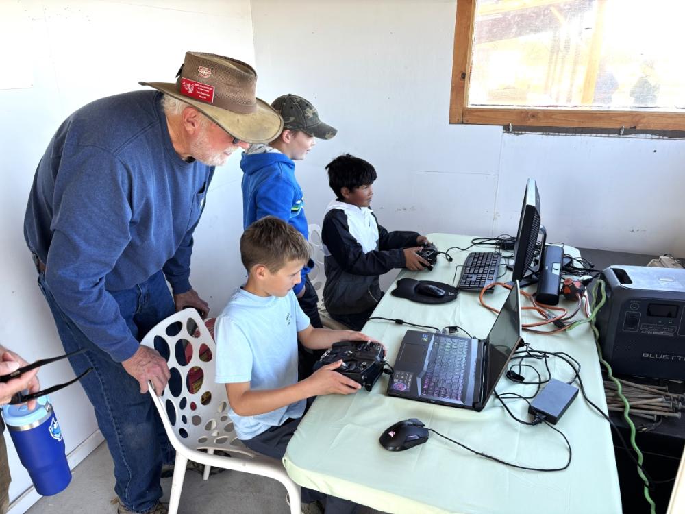 Older man and three boys using laptops and gaming controllers at a table.