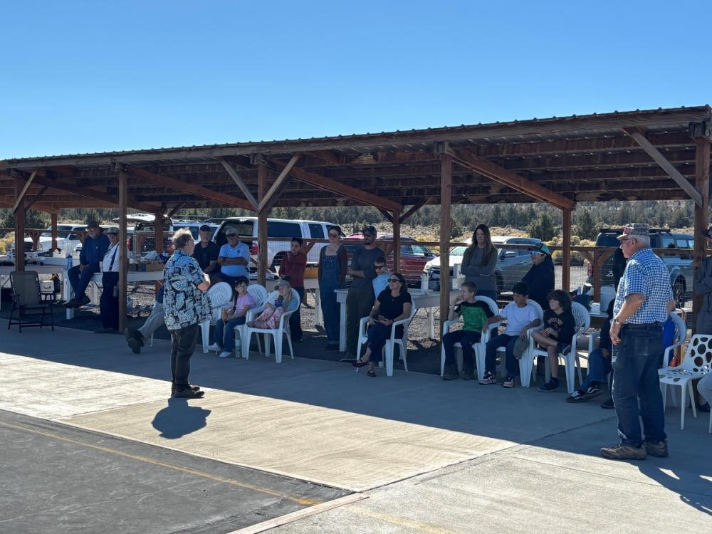 People sitting and standing under a shaded pavilion on a sunny day.