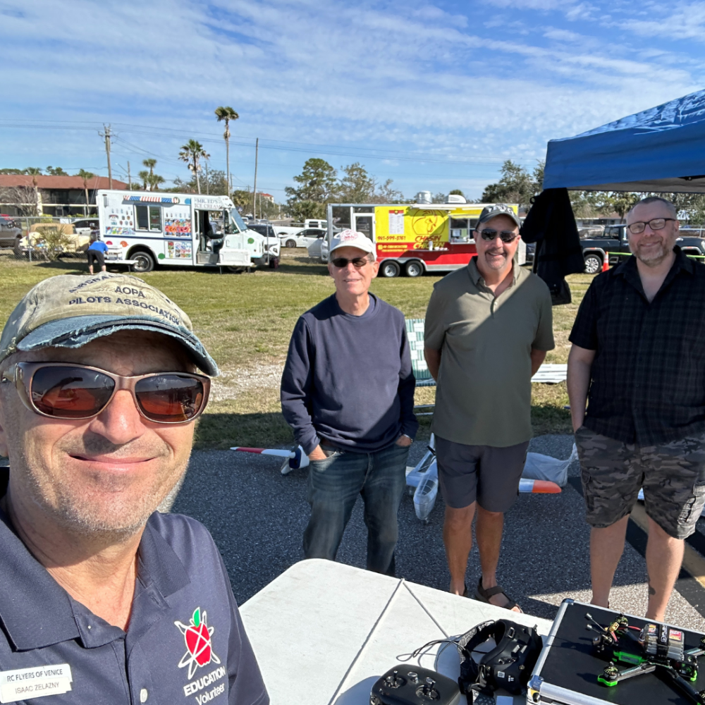 Four men smiling outdoors near food trucks and a tent on a sunny day.