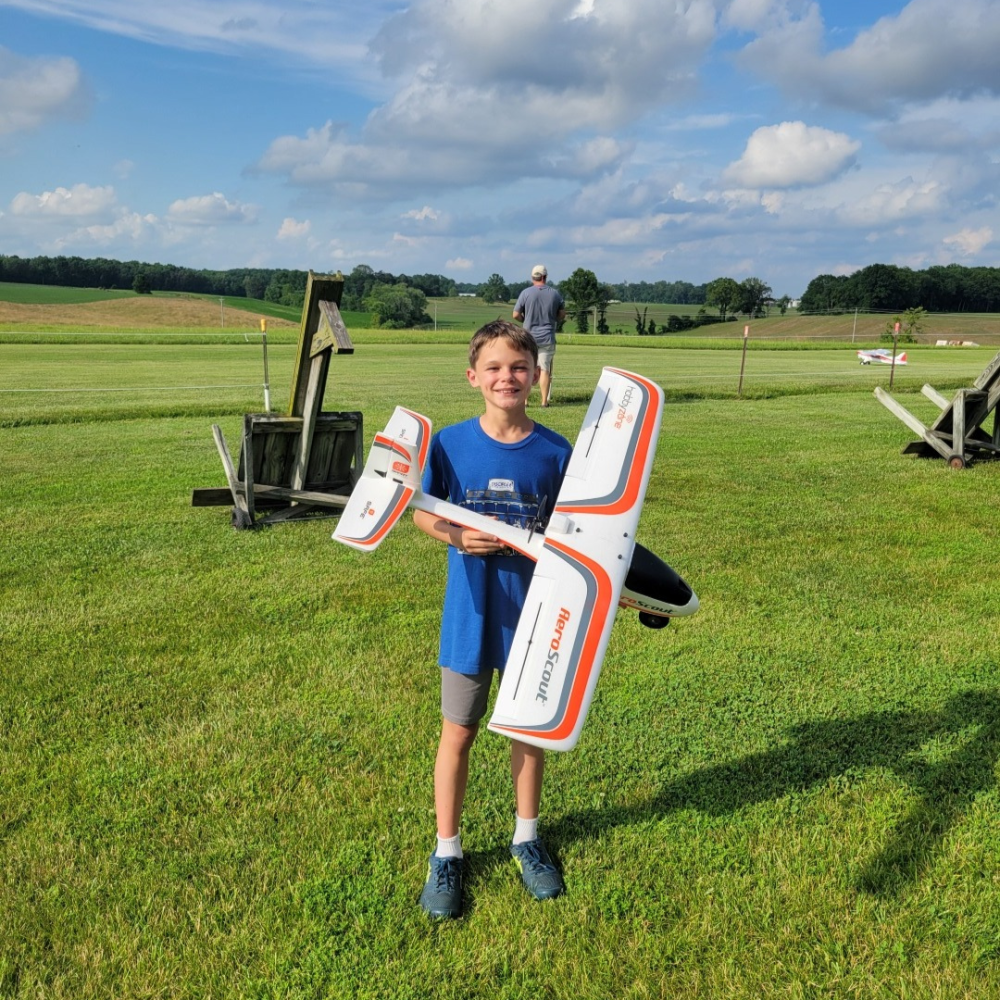 Child holding a model airplane on a grassy field, with a clear blue sky.