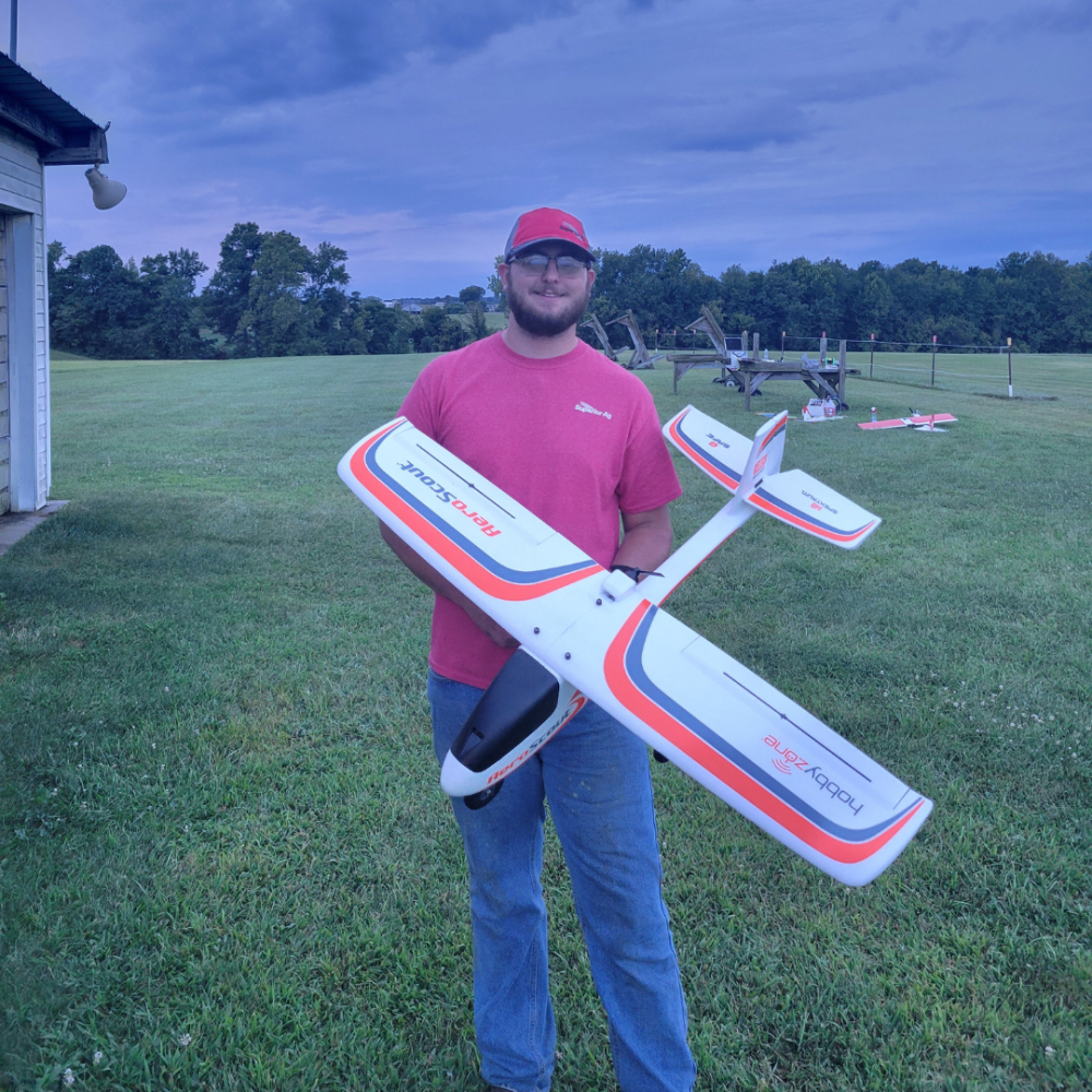 Man in a red shirt holding a model airplane on a grassy field.