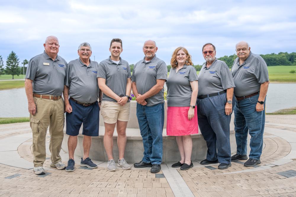 Seven people stand together, smiling, in front of a fountain in a park.