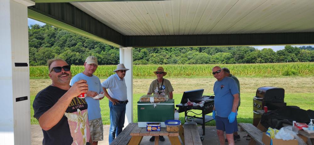 Group of six men socializing under a pavilion, one holding a soda can, with a green landscape view.
