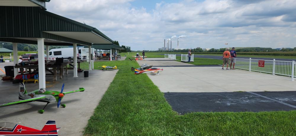Airfield with model planes, people standing nearby, runway, and green field under a cloudy sky.