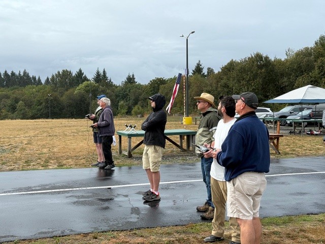 Five people observing RC planes on a grassy field, American flag in background.