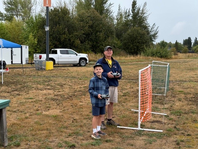 Two people in a grassy field with remote controls, next to a white pickup truck.
