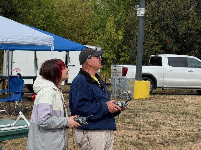 Two people using remote controls outdoors, with tents and a truck nearby.