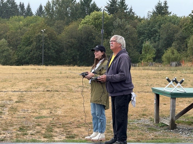 Two people flying drones in a park, trees in the background.