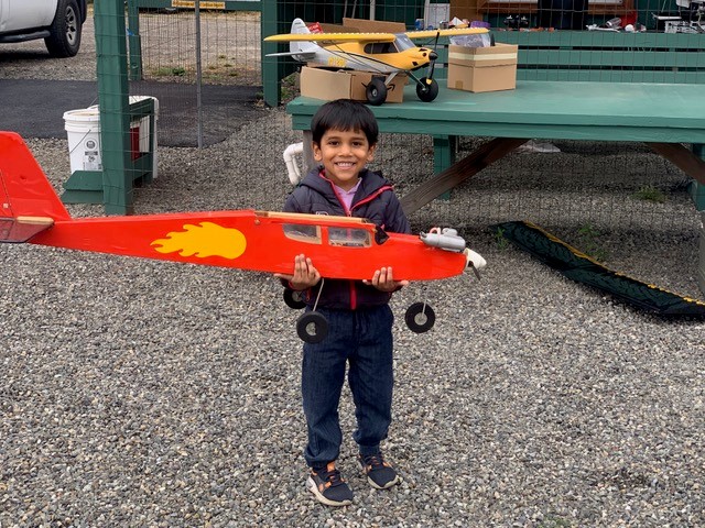 Child smiling, holding a red model airplane outdoors on gravel.