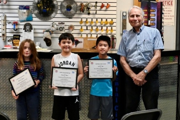 Three children holding certificates, standing beside an adult in a store setting.