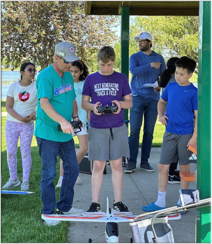 Group flying model planes at a park, young boy operating a remote control.
