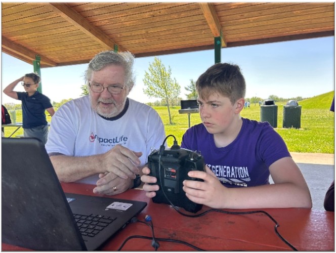 A man and a boy focus on a laptop outdoors under a pavilion.