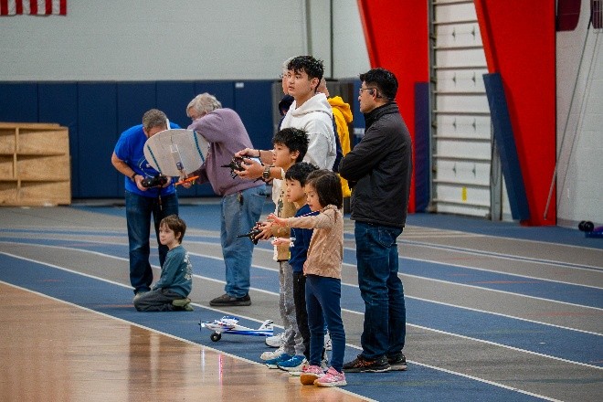 Adults and children control model planes in an indoor gym.