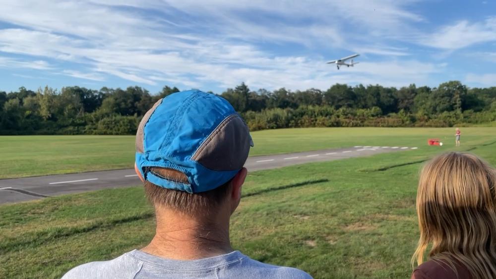 Two people watch a small plane flying over a grassy field on a sunny day.