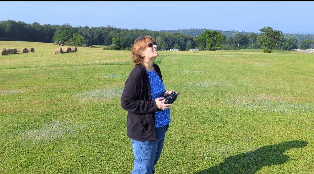 Woman smiling, flying a drone in a green field under clear blue sky.