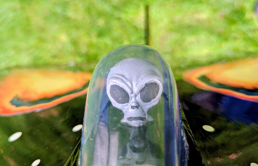 Alien face model inside a transparent dome, against a colorful and blurred background.