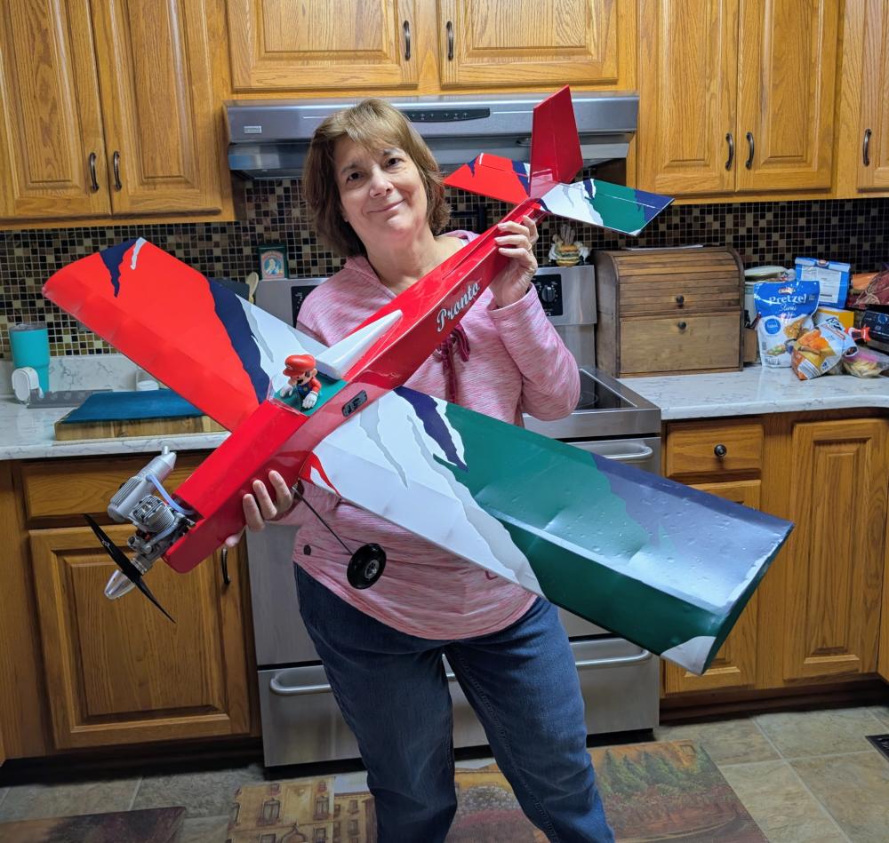 Woman smiling, holding a red model airplane in kitchen.