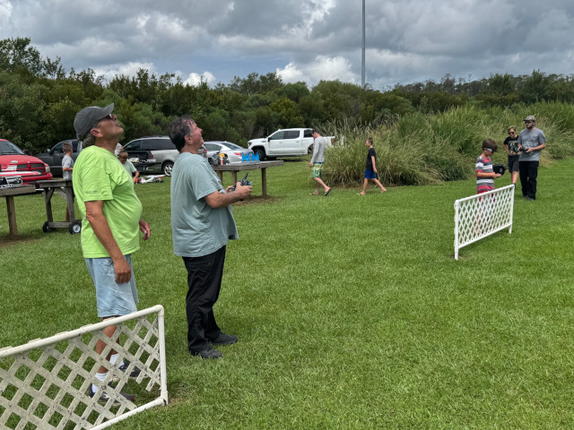 Two men flying drones in a grassy field with people in the background.