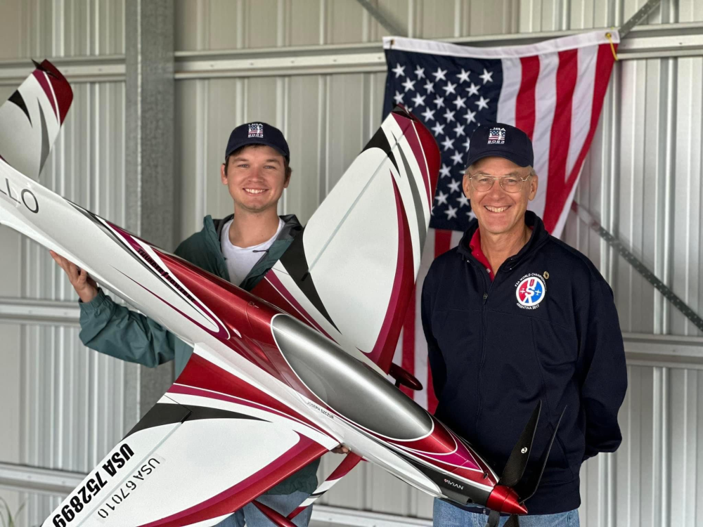 Two people smiling, holding a large model airplane in front of a U.S. flag.