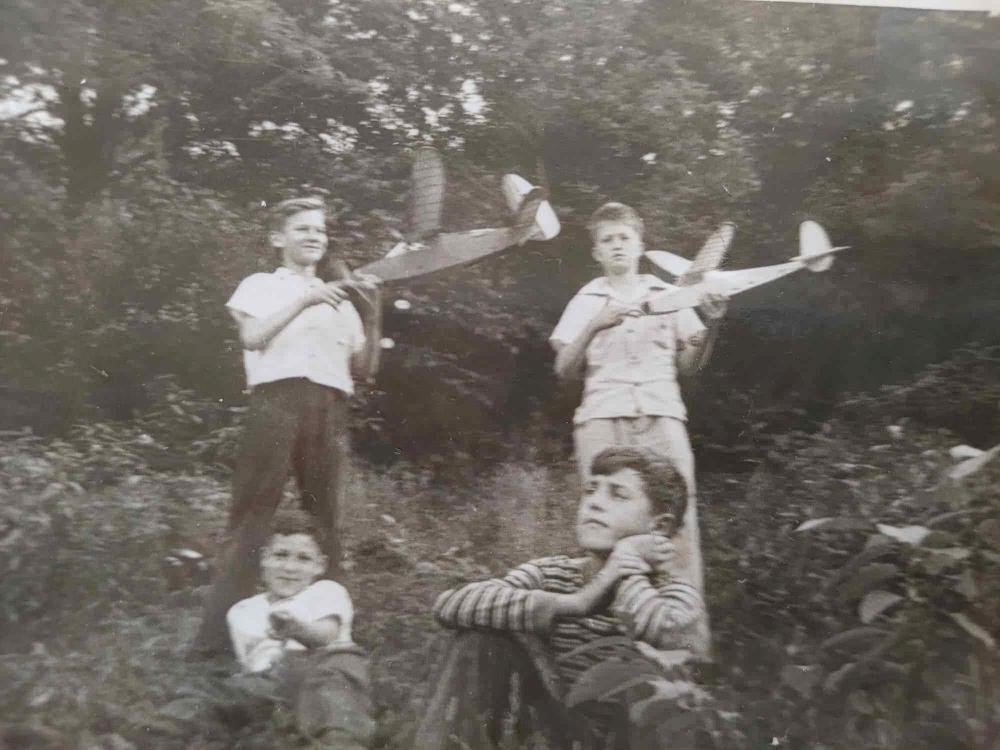 Black-and-white photo of boys holding model planes outdoors, with two sitting on ground.