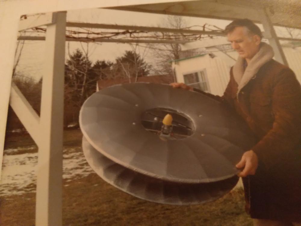 Man holding a large metallic disc outdoors, wearing a brown coat.