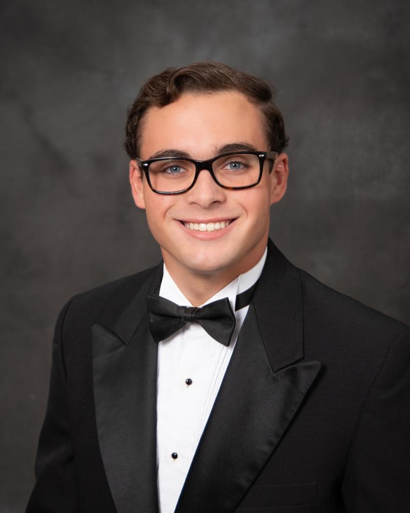 Young man in a tuxedo and glasses, smiling against a dark backdrop.