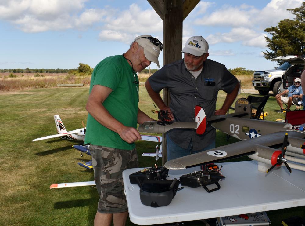 Two men examine model airplanes on a table outdoors.