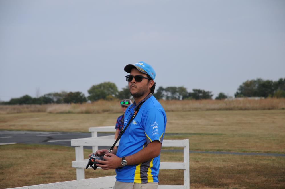 Man in blue shirt and cap stands on grassy field holding a remote control.