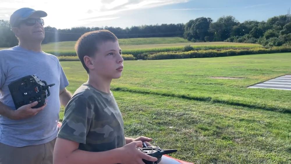 Boy and man operating remote-controlled planes in a grassy field under a blue sky.