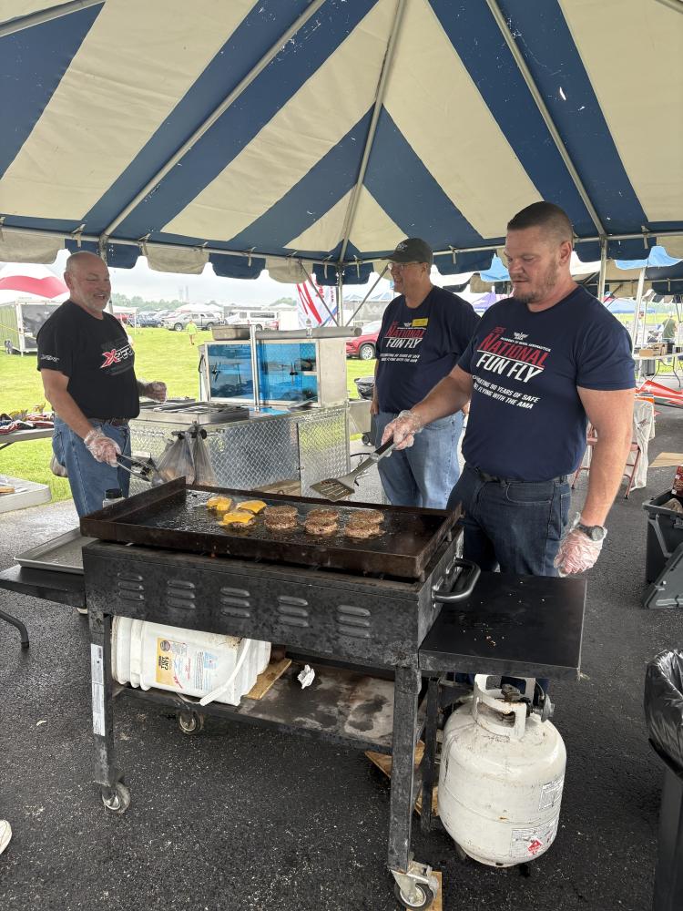 Three men grilling burgers under a striped canopy at an outdoor event.
