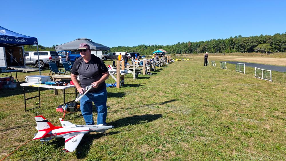 Man preparing a model airplane on a grassy field with tents and people in the background.