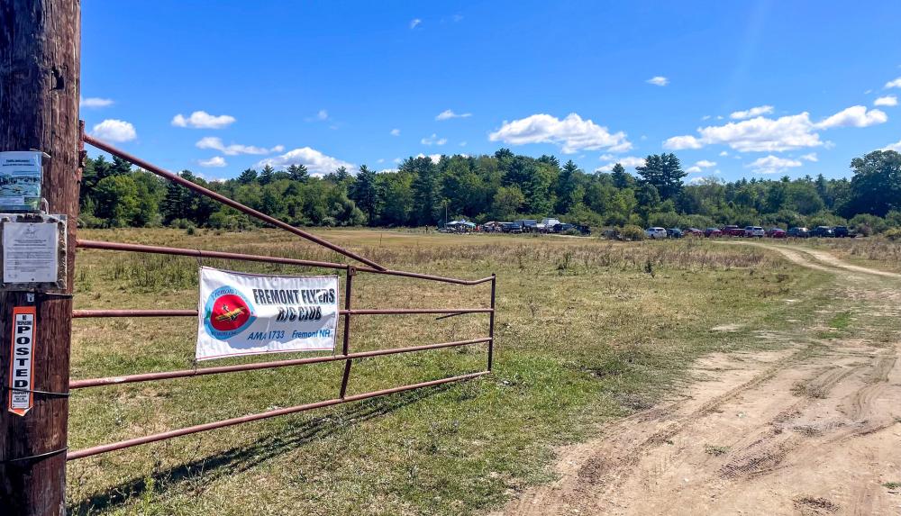 Dirt road leading to an open field with a metal gate and banner under a clear blue sky.