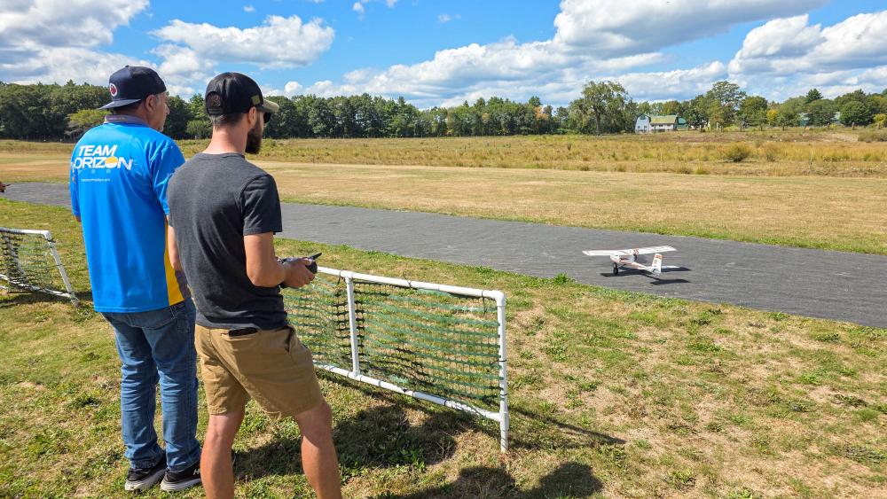 Two men piloting a model plane on a grassy airfield under a partly cloudy sky.
