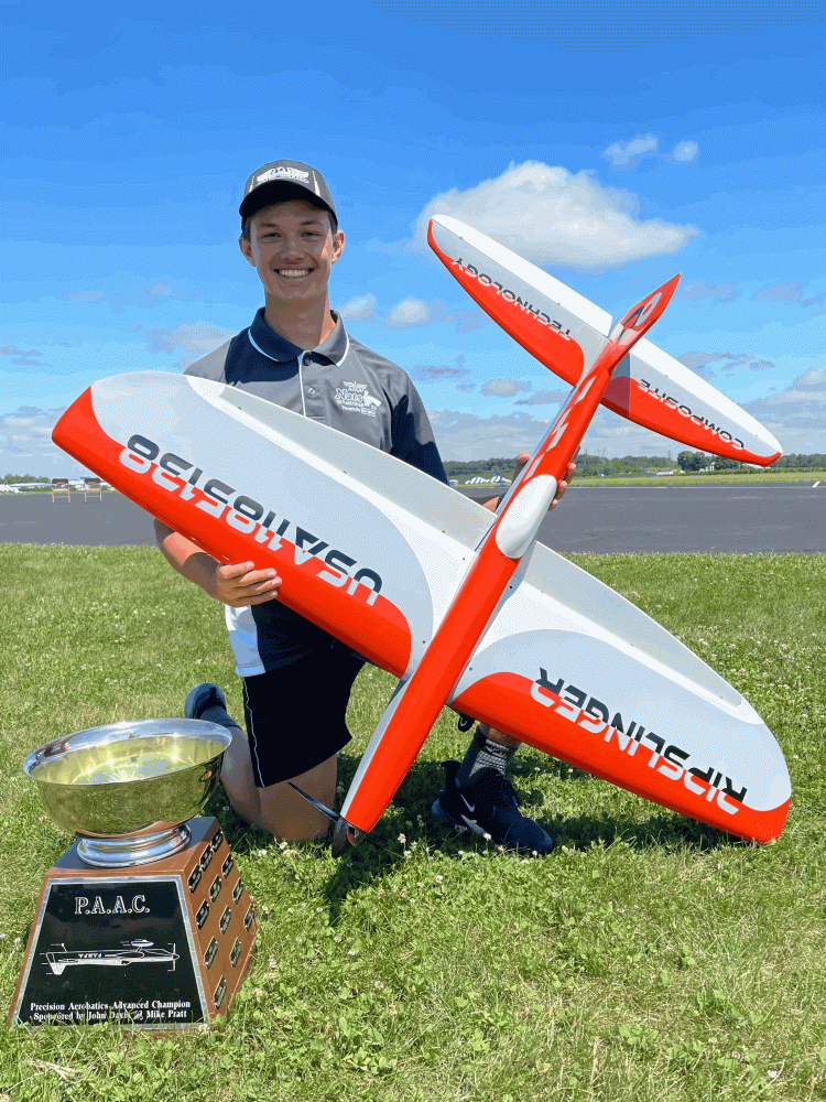 Person kneels on grass, holding a red and white model plane with a trophy nearby.