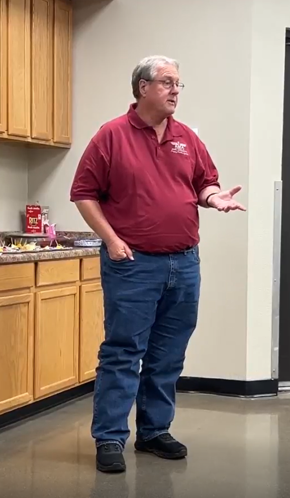 Man in red shirt and jeans speaking, stands by wooden cabinets and snacks.