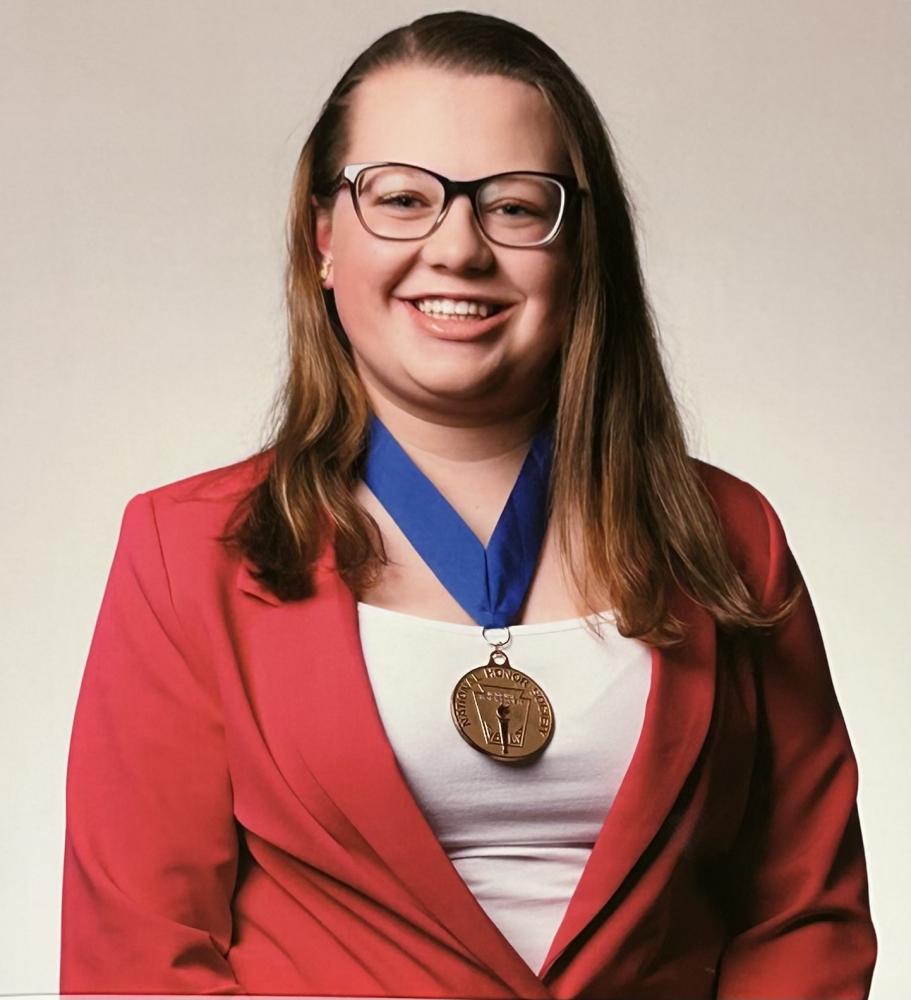 A person smiling, wearing glasses, a red blazer, and a medal on a blue ribbon.