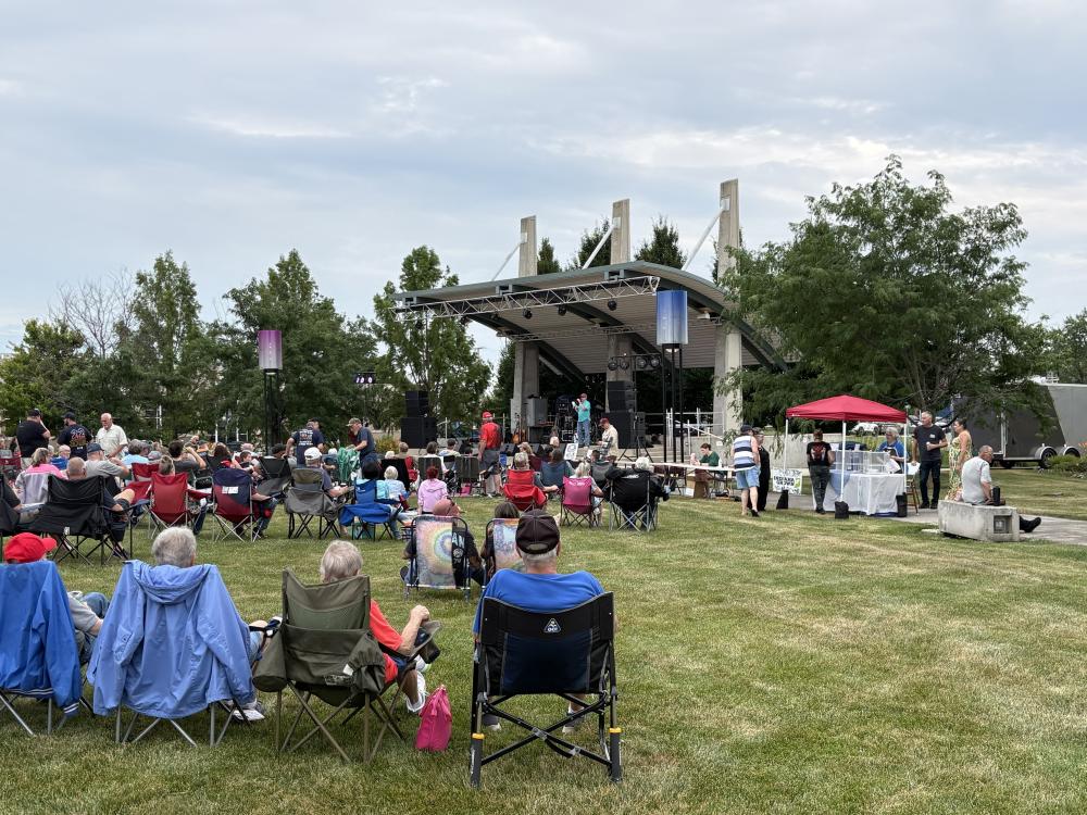 Outdoor concert with seated crowd facing a stage under a cloudy sky.